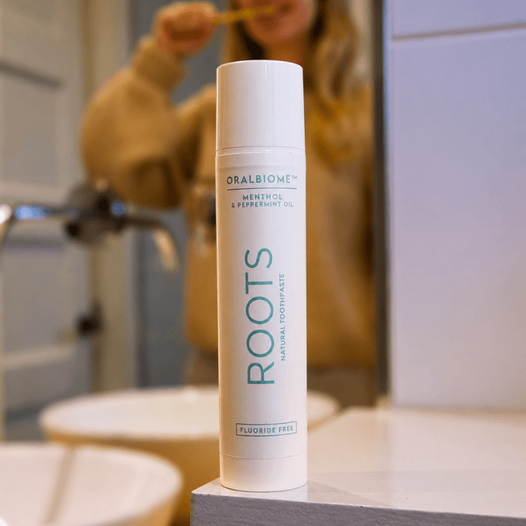 A tube of Oralbiome Roots toothpaste on a bathroom counter with a person brushing their teeth in the background.