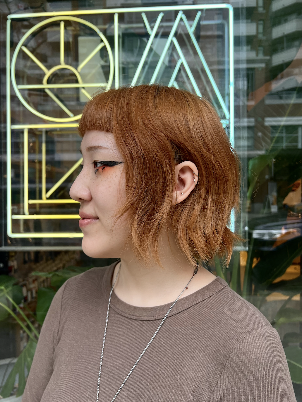 Person with short red hair and eyeliner, standing in front of a neon geometric sign.