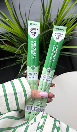 Two green organic meat stick packages held in front of a plant.