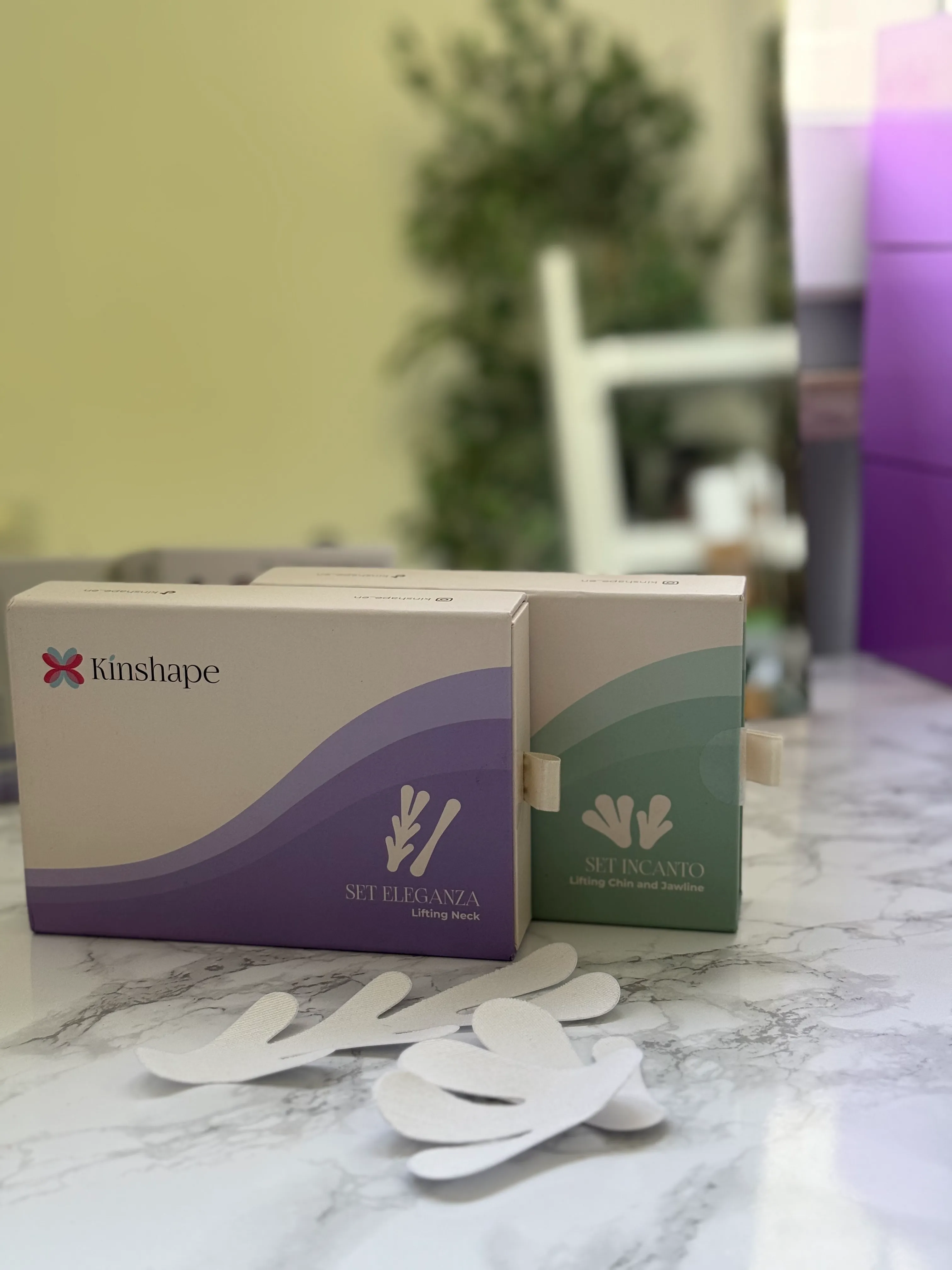 Two Kinshape product boxes for neck and chin lifting sit on a marble surface with paper cutouts.