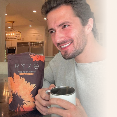 Person smiling with a mug, next to a bag of Ryze mushroom coffee.