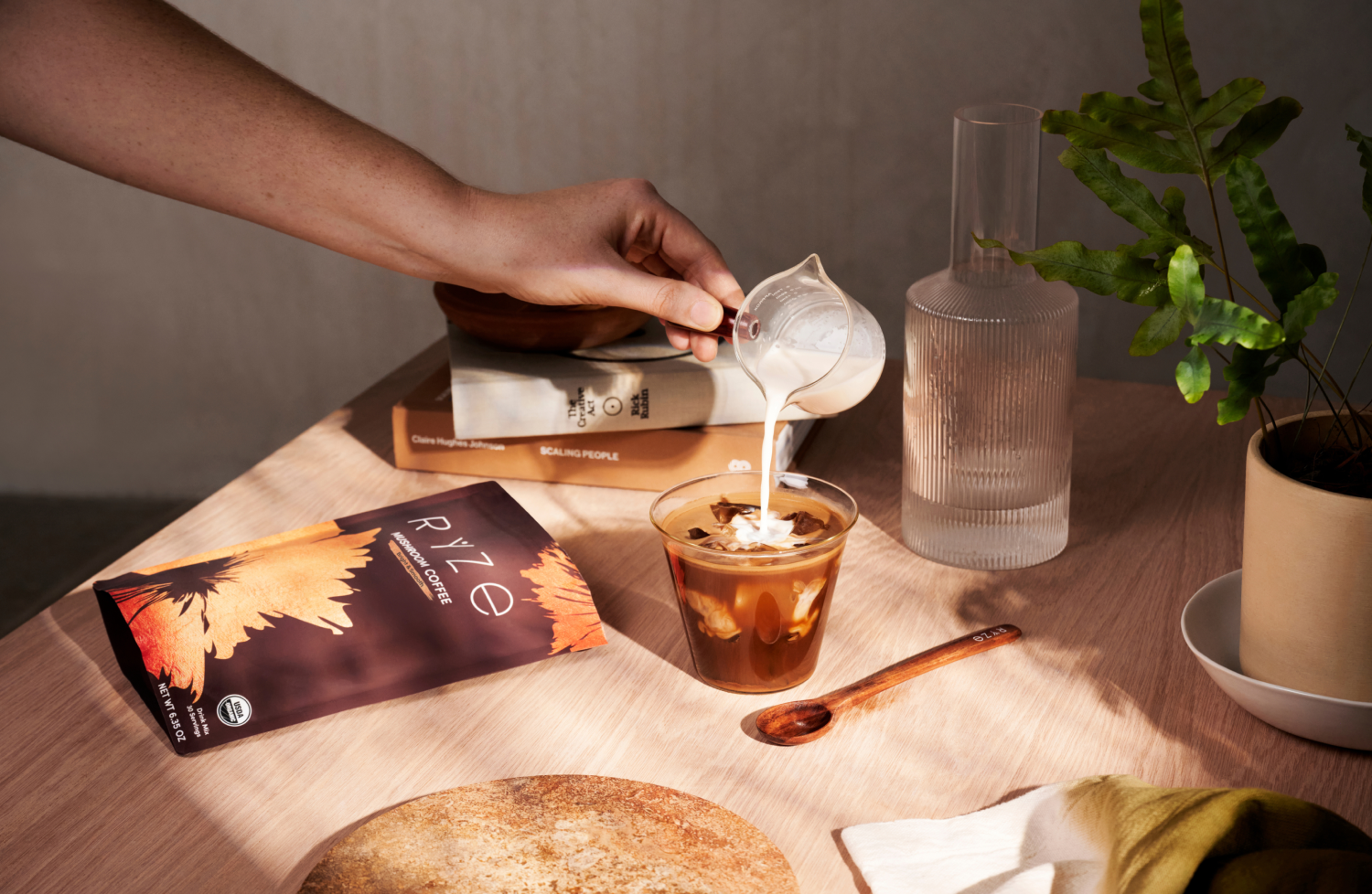 A hand pouring milk into iced coffee next to books, a plant, and a Ryze Coffee package.