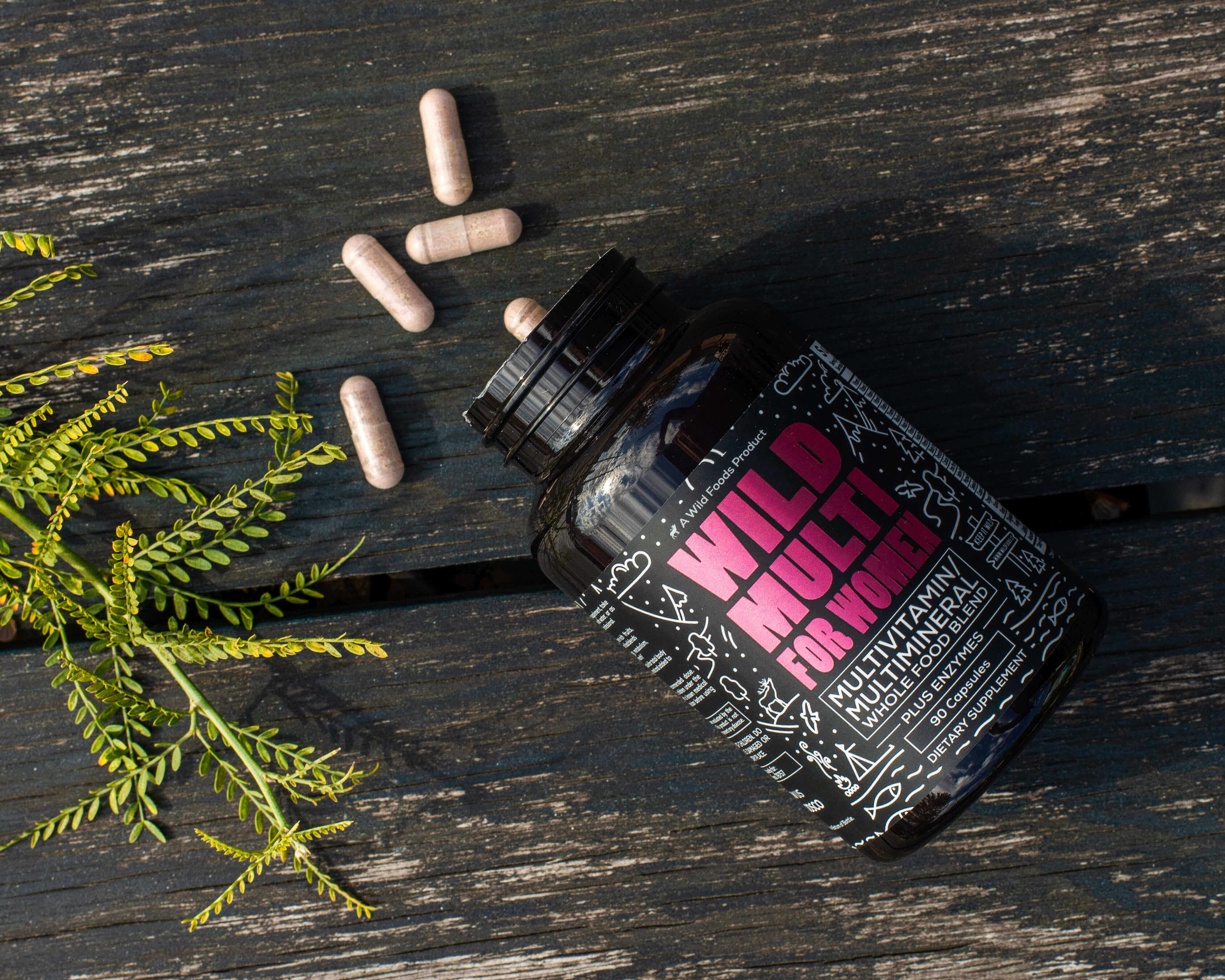 Multivitamin bottle labeled 'Wild Multi for Women' with capsules and green leaves on wooden surface.