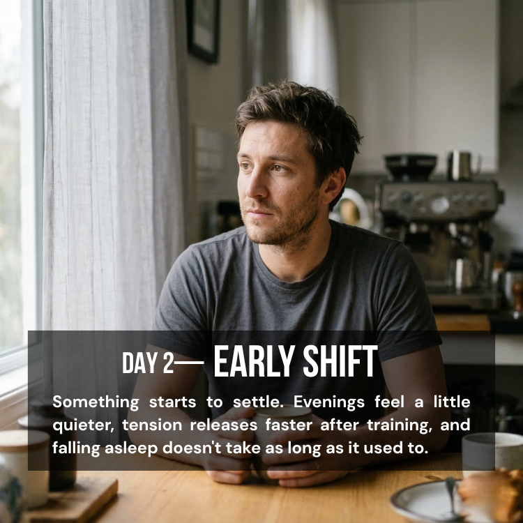A man in a grey t-shirt sits at a table and looks thoughtfully out of a window.