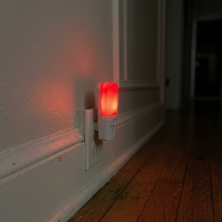 A small, red night light plugged into a wall outlet, illuminating a dark hallway.