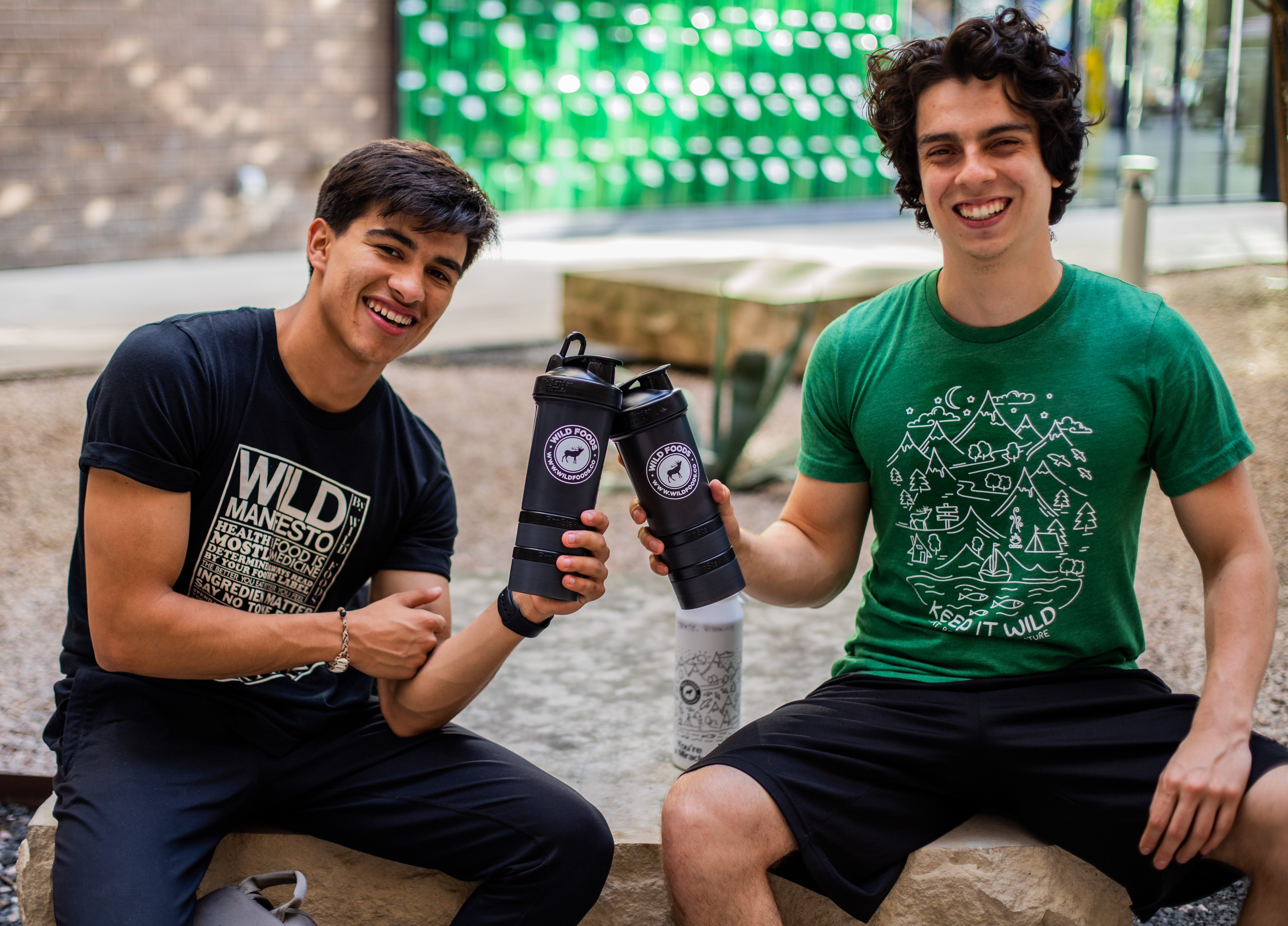 Two people sitting outdoors, smiling and holding black bottles, wearing printed t-shirts.