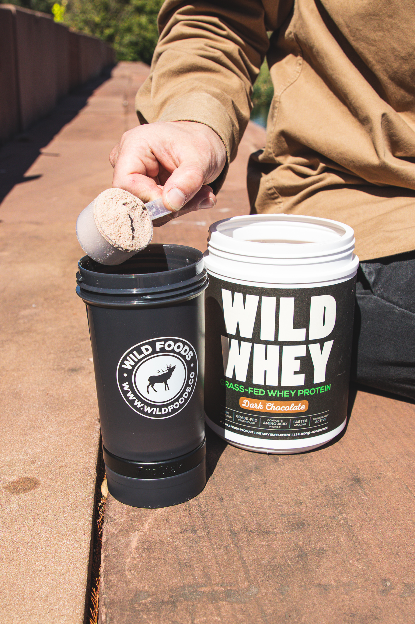 Person scooping whey protein into a shaker bottle beside the container.