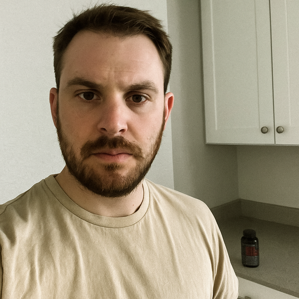 A person with a beard standing in a kitchen with white cabinets and a bottle on the counter.