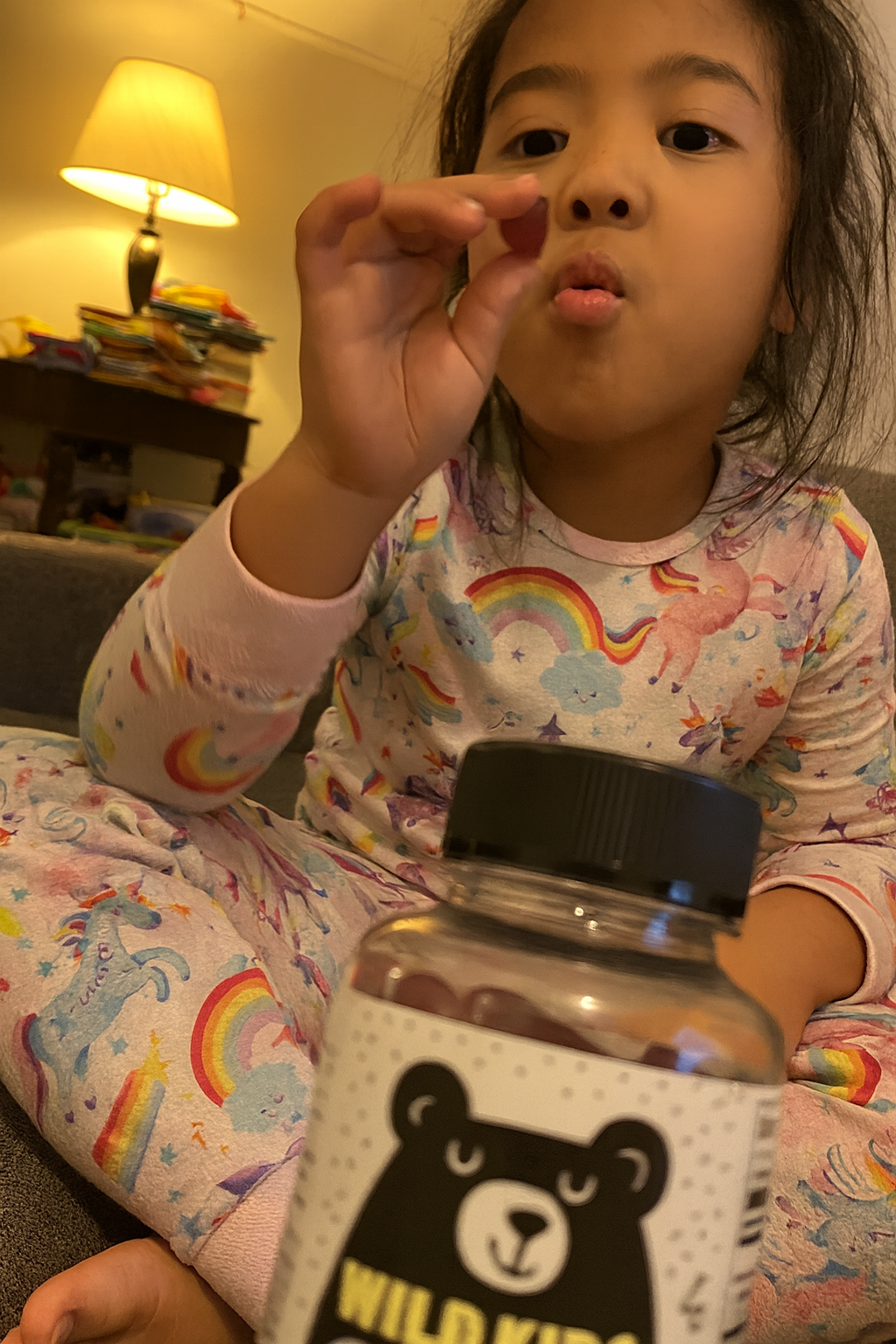 Child in pajamas holding a gummy near a jar, with a lamp in the background.