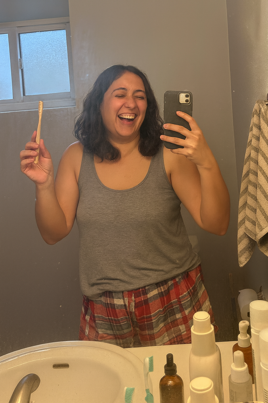 Person in mirror holding toothbrush and phone, smiling in a bathroom.