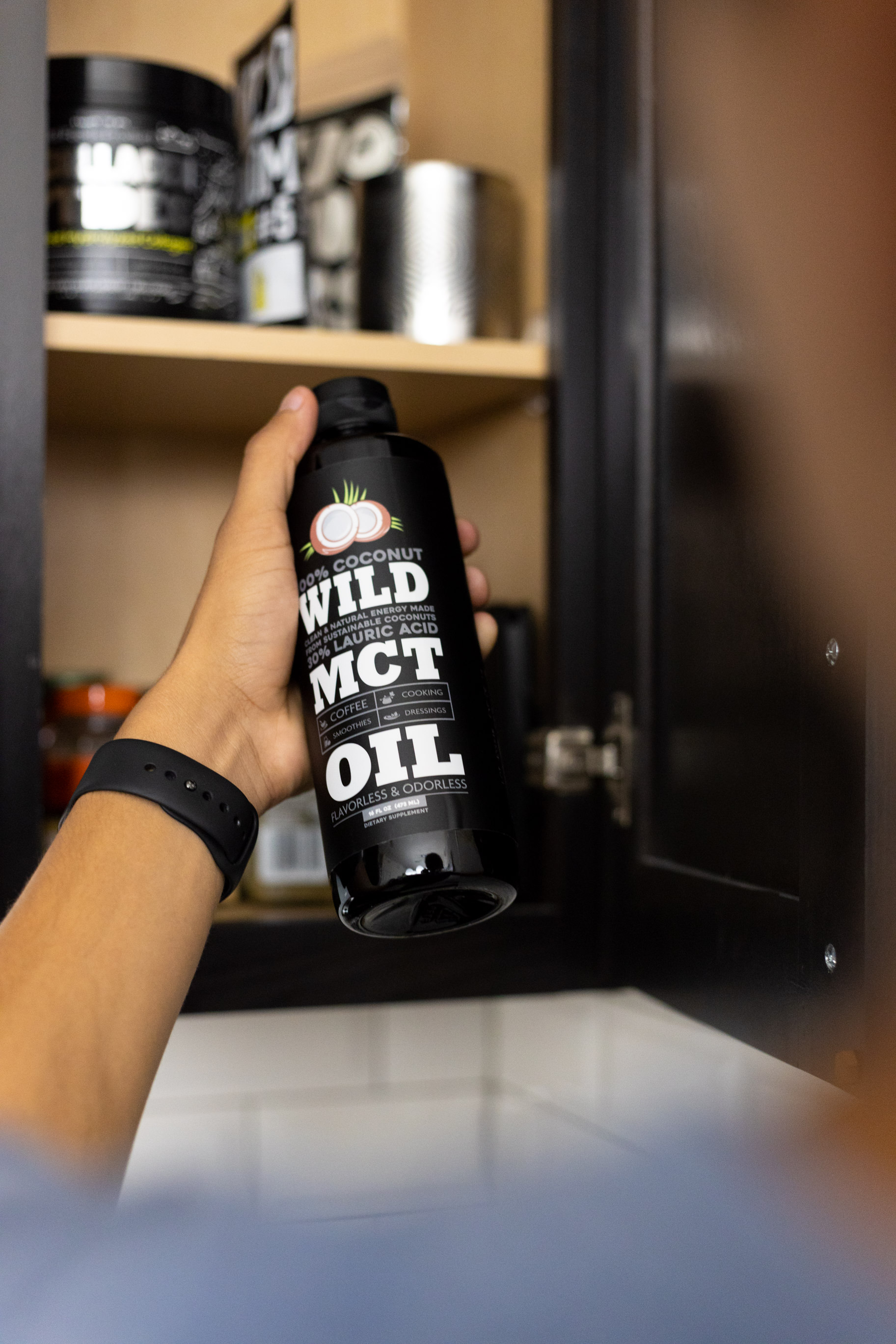 Hand holding a bottle of Wild MCT oil in front of an open cabinet.