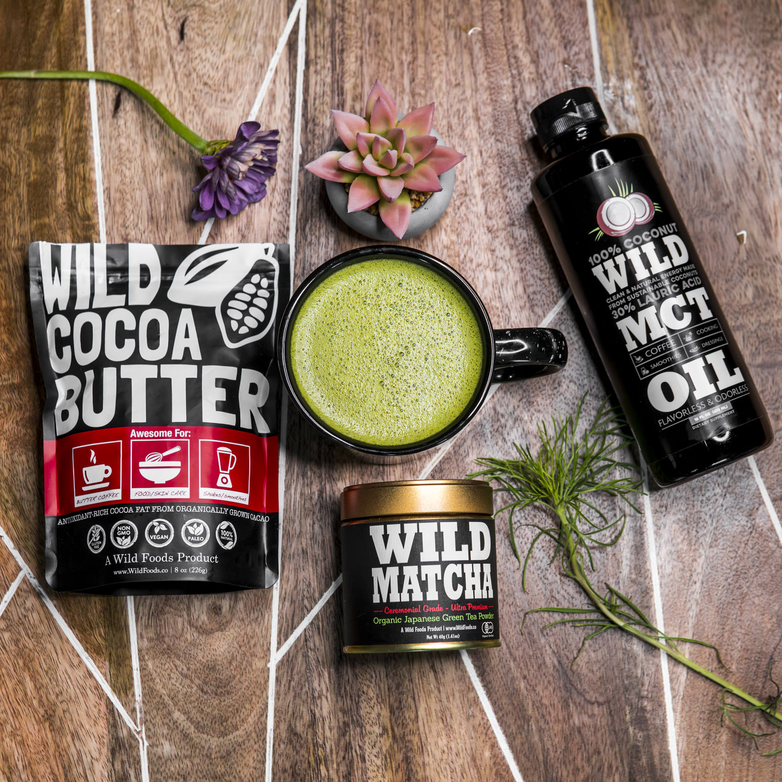 Assorted products on wooden table: cocoa butter, matcha, MCT oil, cup with green drink, flowers.