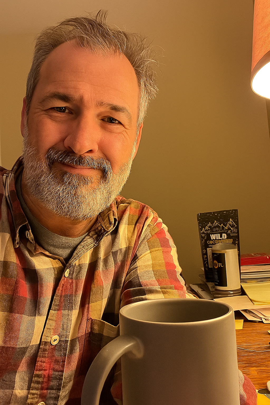 Person smiling with a gray mug in front, wearing a plaid shirt indoors.