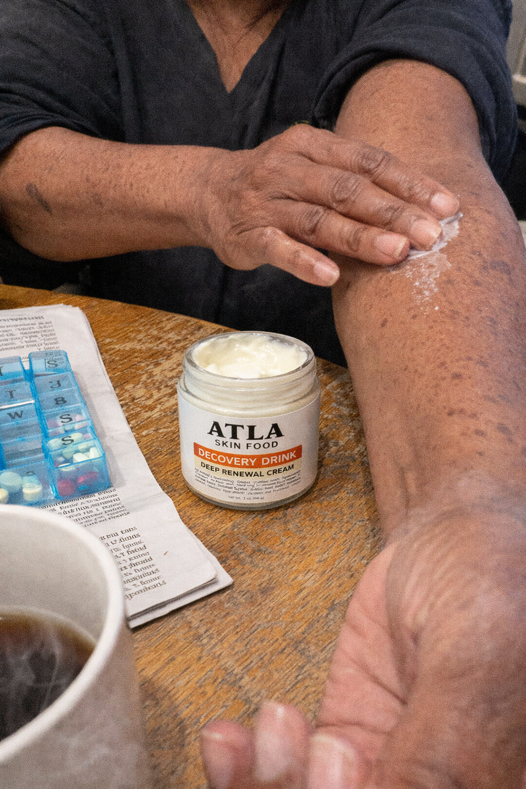 A person applies white cream from a jar labeled 'ATLA SKIN FOOD' onto their arm at a table.