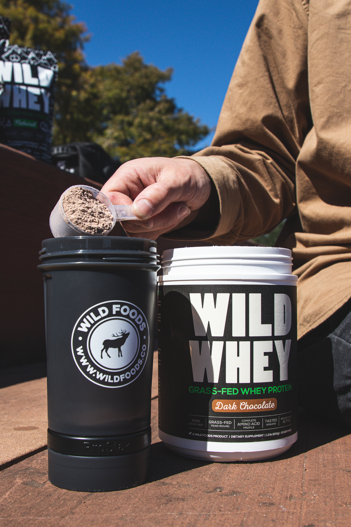 Person scooping Wild Whey protein powder into a shaker bottle outdoors.