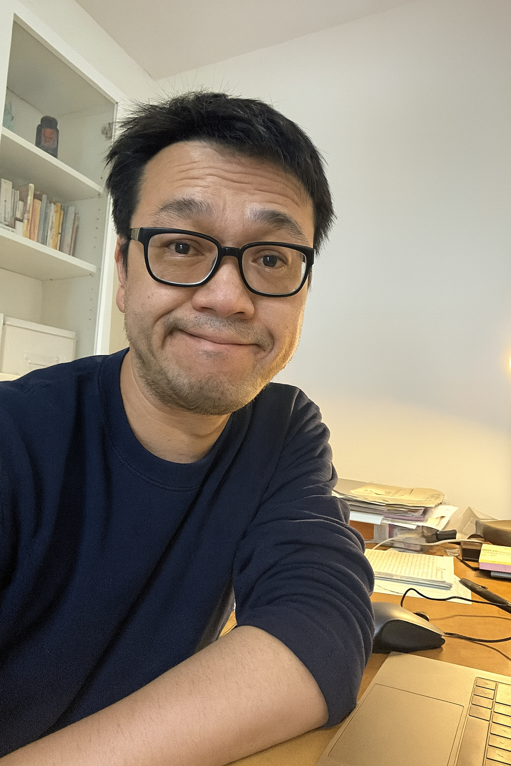 Person wearing glasses sitting at a desk with books and papers in the background.