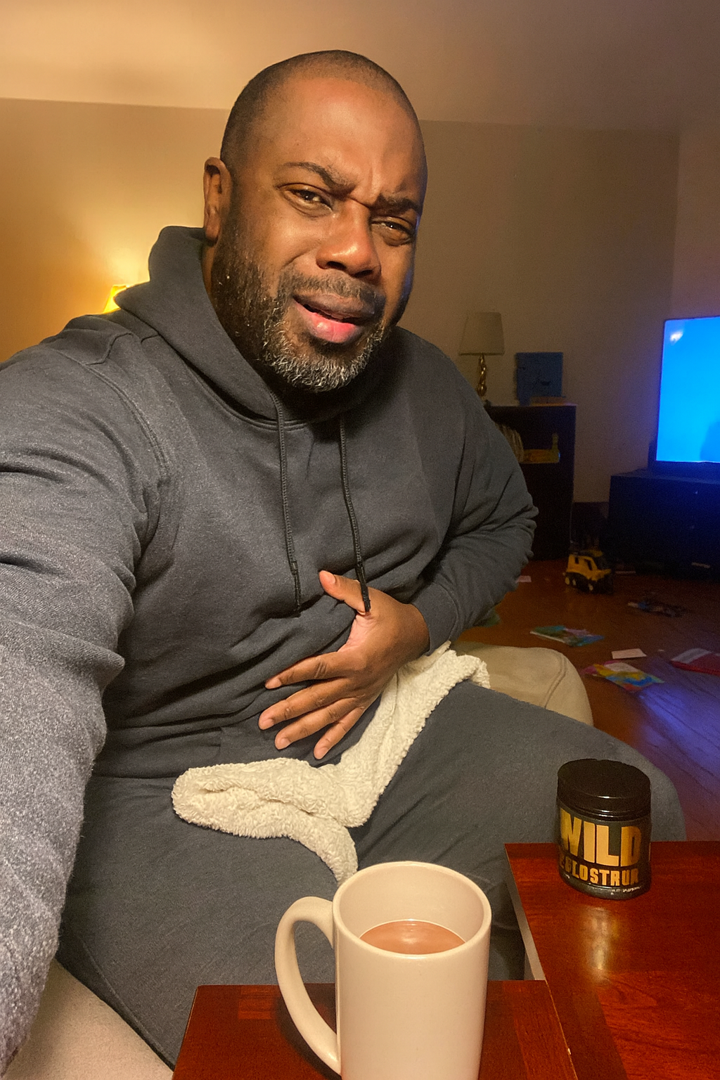 Person in hoodie sitting with a mug and holding stomach, appearing uncomfortable.