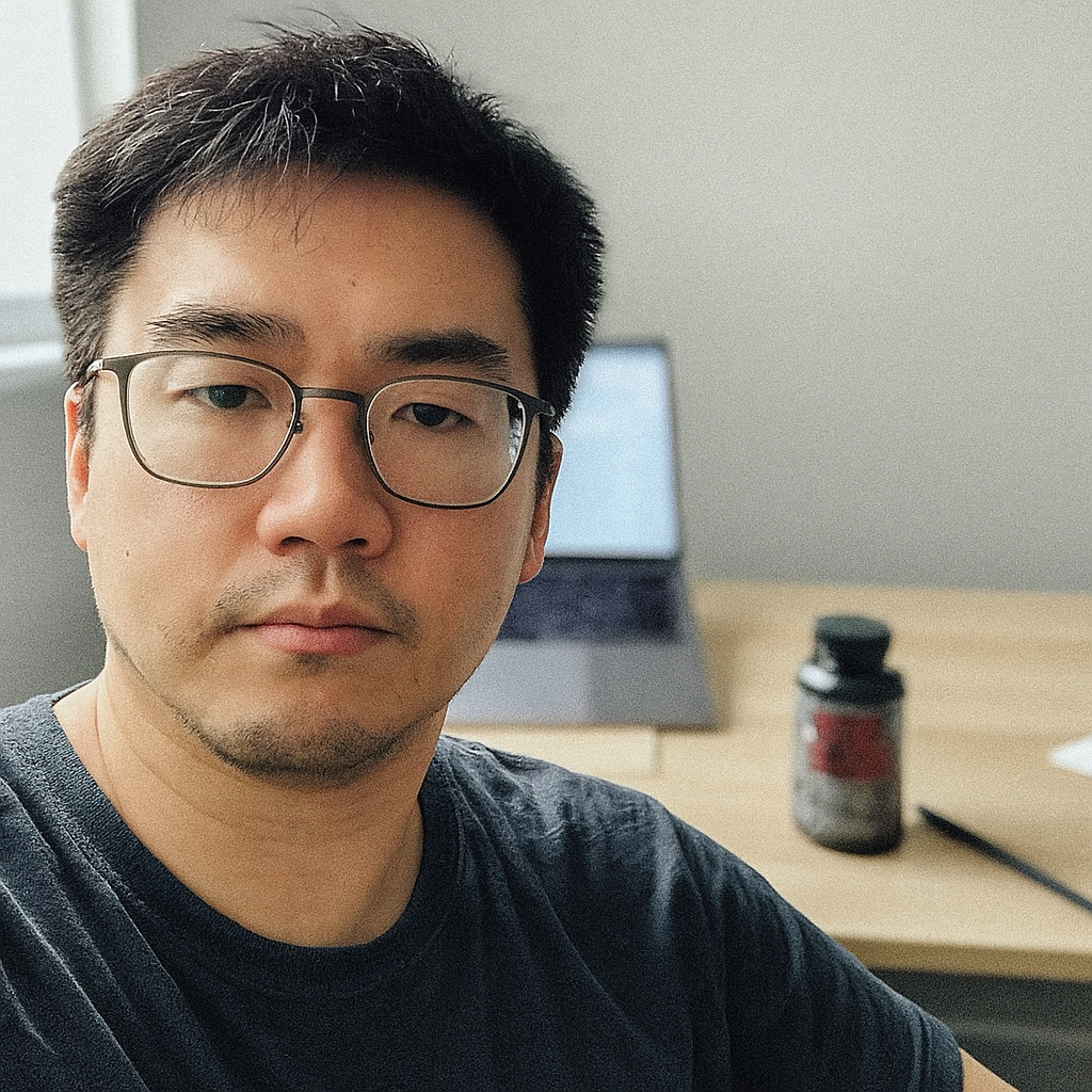 Person with glasses sitting in front of a desk with a laptop and bottle.