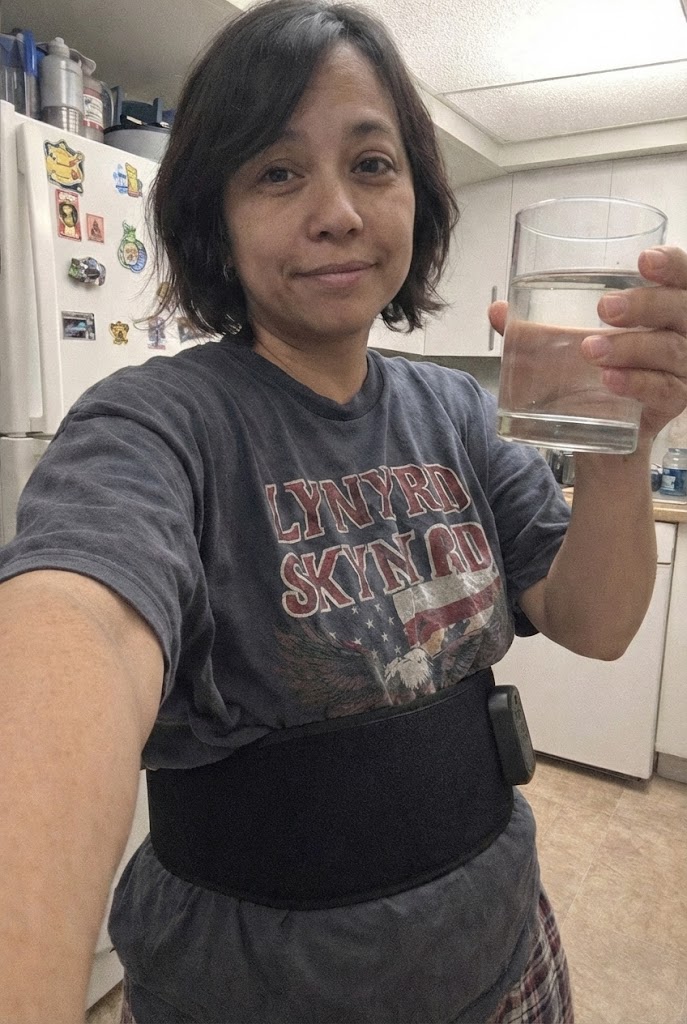 A woman wearing a Lynyrd Skynyrd t-shirt and a black waist belt holds a glass of water.