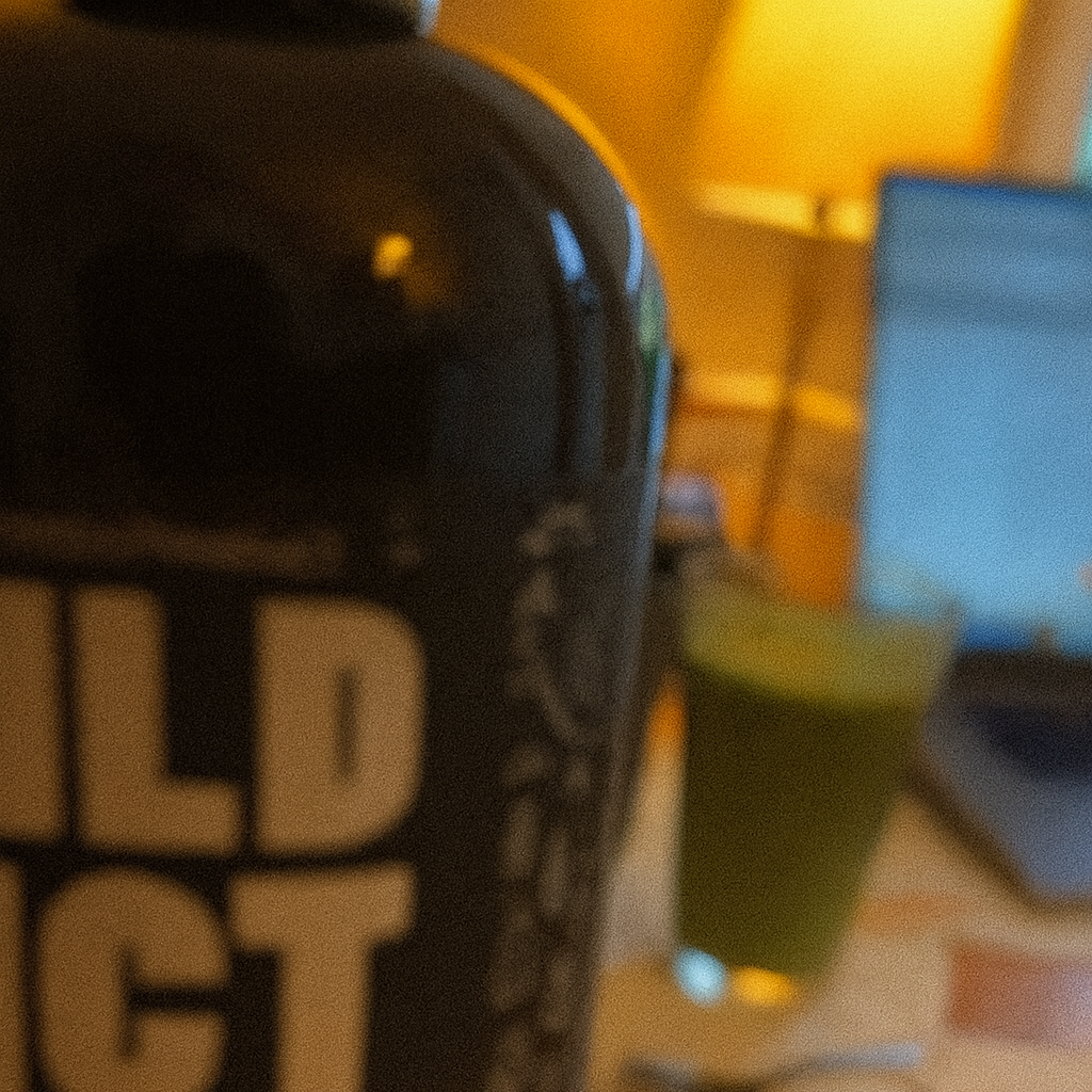 Close-up of a black bottle with partial text, blurred background with laptop and lamp.