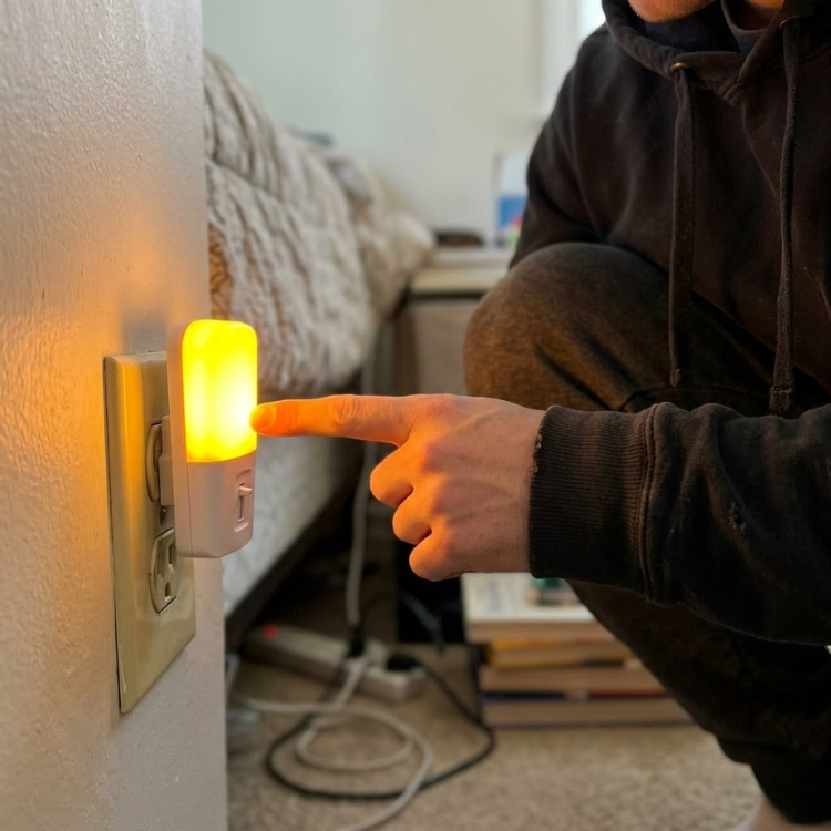 A person's finger touches a glowing yellow nightlight plugged into a wall outlet in a bedroom.