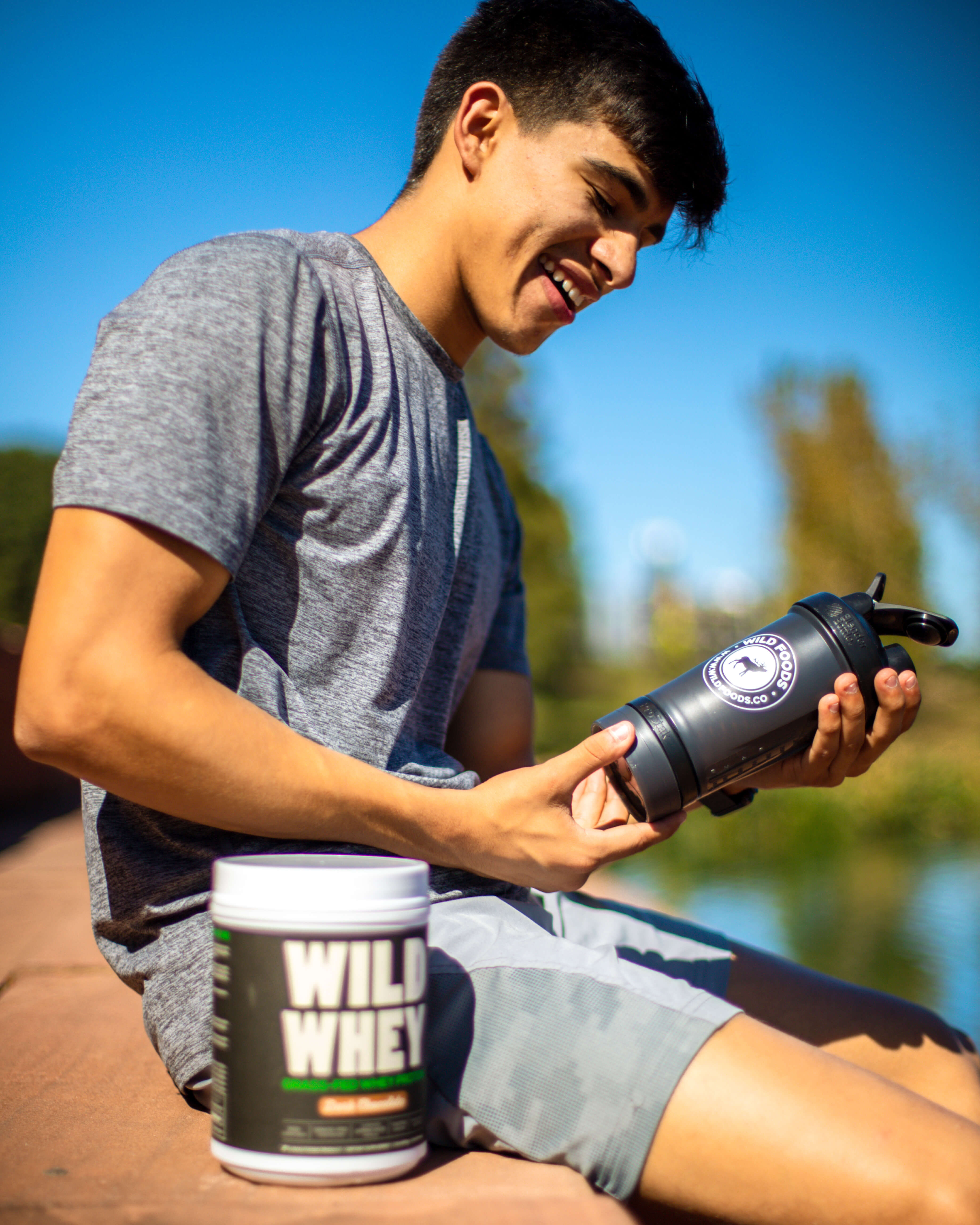Person sitting outdoors holding a shaker bottle near a container labeled 'Wild Whey'.