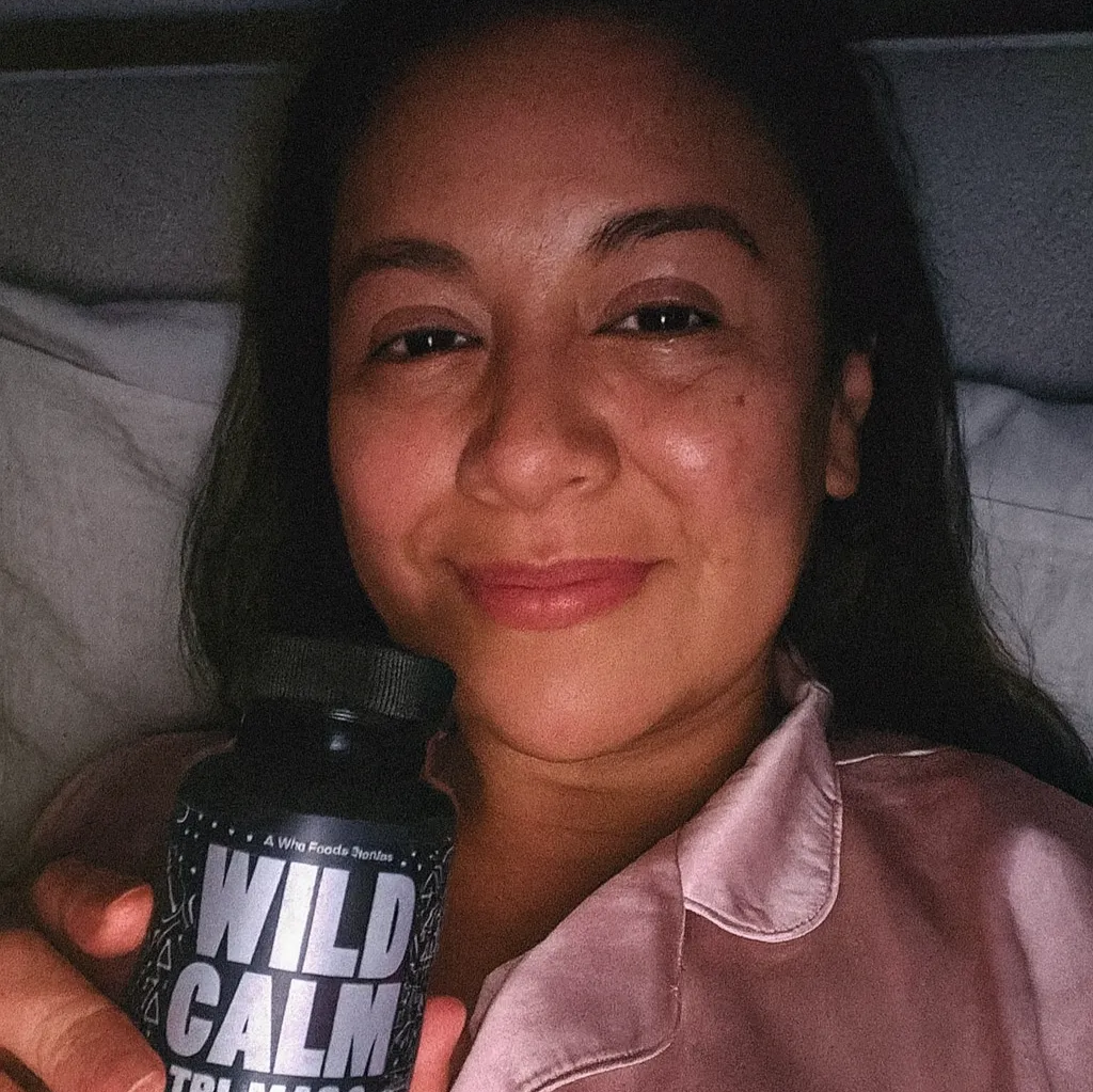 Person smiling and holding a bottle labeled 'Wild Calm' in bed.