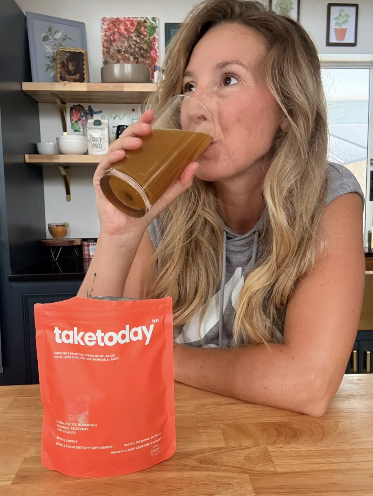 A woman with blonde hair drinks from a glass next to a red pouch of Taketoday supplement.