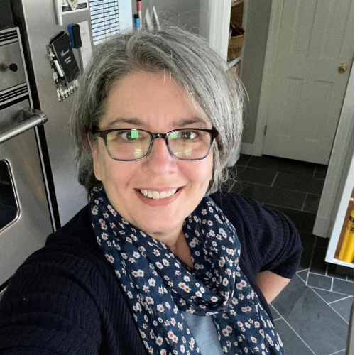 A smiling woman with short gray hair and glasses, wearing a blue floral scarf, takes a selfie.