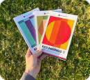 A hand holding three colorful, geometrically designed booklets over a sunlit grassy lawn.