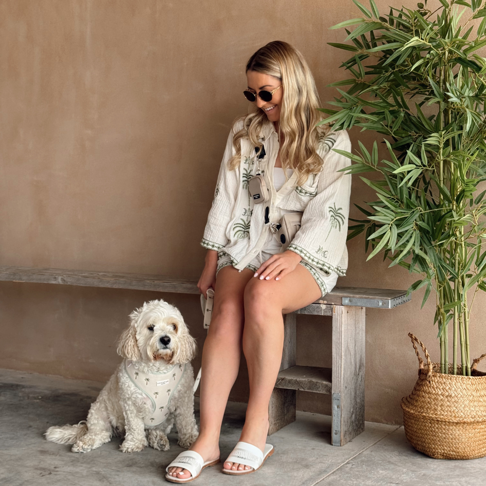 A woman sits on a wooden bench smiling at her small white dog sitting on the floor beside her.