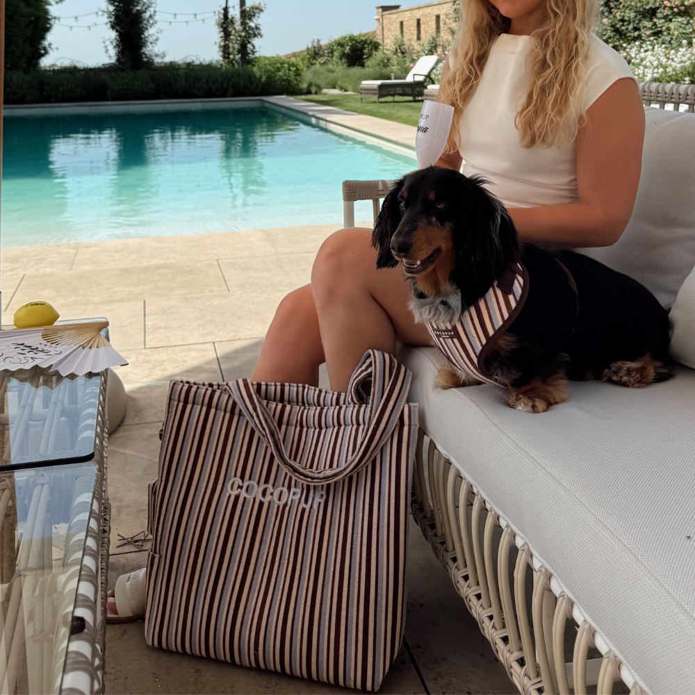 A woman and a dachshund sit on an outdoor sofa by a pool, with a striped tote bag nearby.