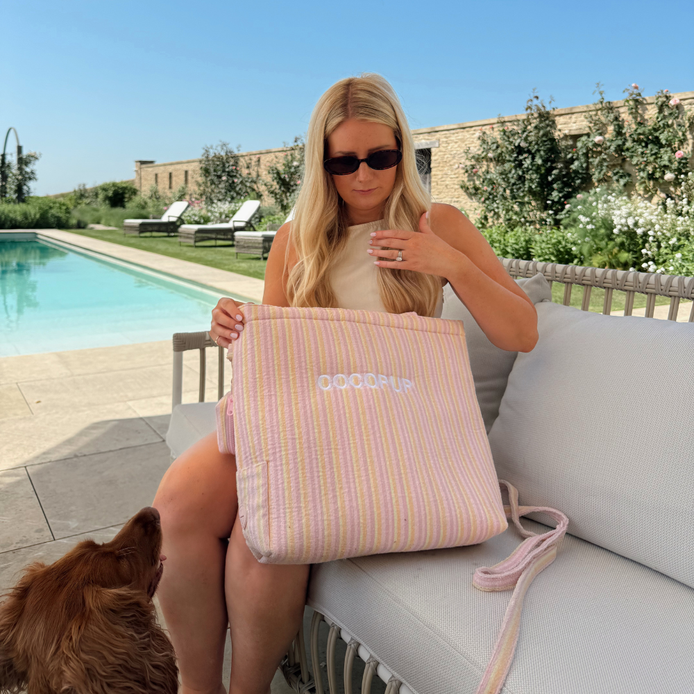 A blonde woman in sunglasses sits by a pool holding a large pink and yellow striped tote bag.