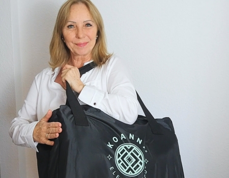 A smiling woman with blonde hair in a white shirt holds a large black bag over her shoulder.