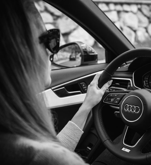 Person driving an Audi, viewed from the passenger side in black and white.