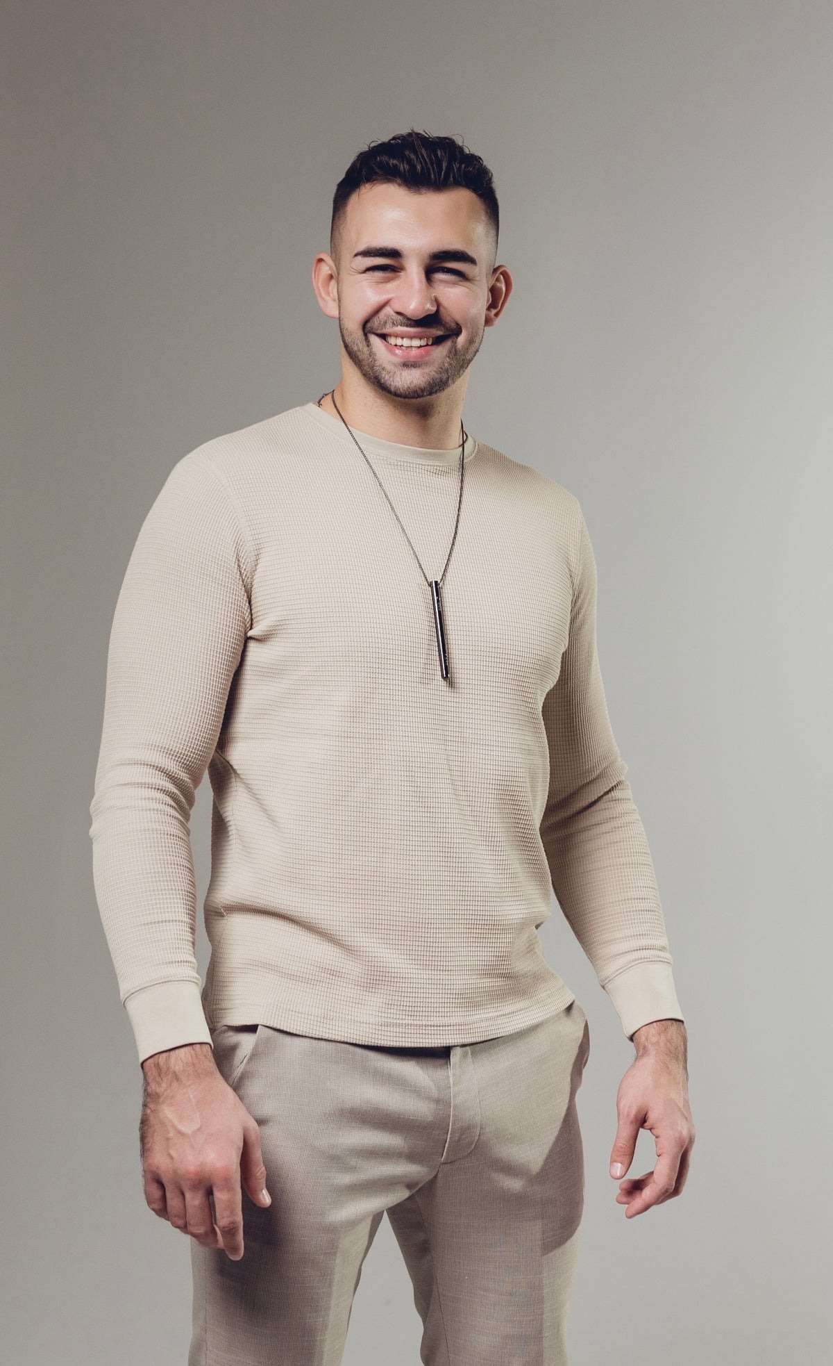 Man smiling, wearing a beige sweater and pants, with neutral background.