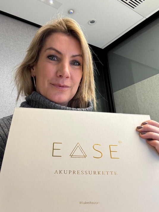 Person holding a box labeled 'EASE Akupressurkette' indoors.