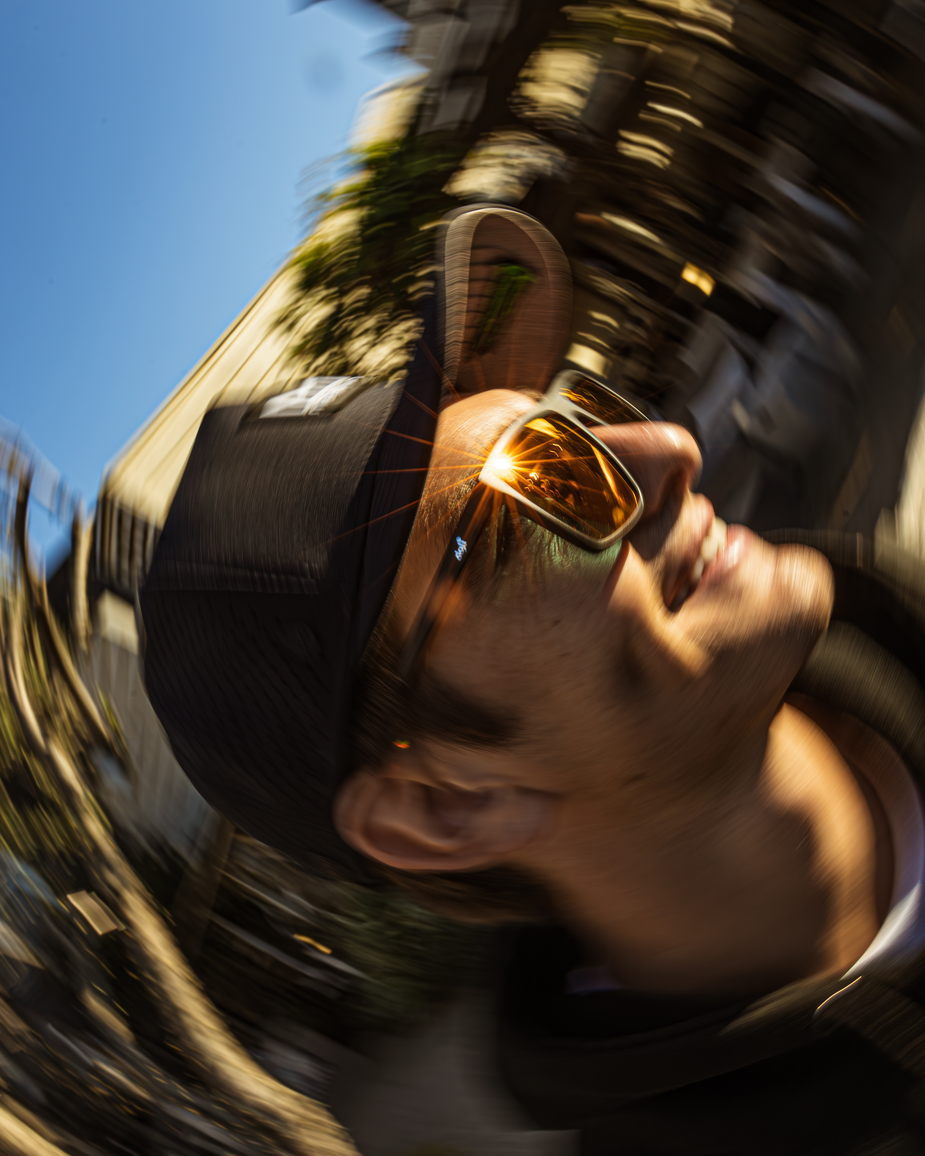 A motion-blurred, low-angle photo of a smiling man in sunglasses with a sun flare on the lens.