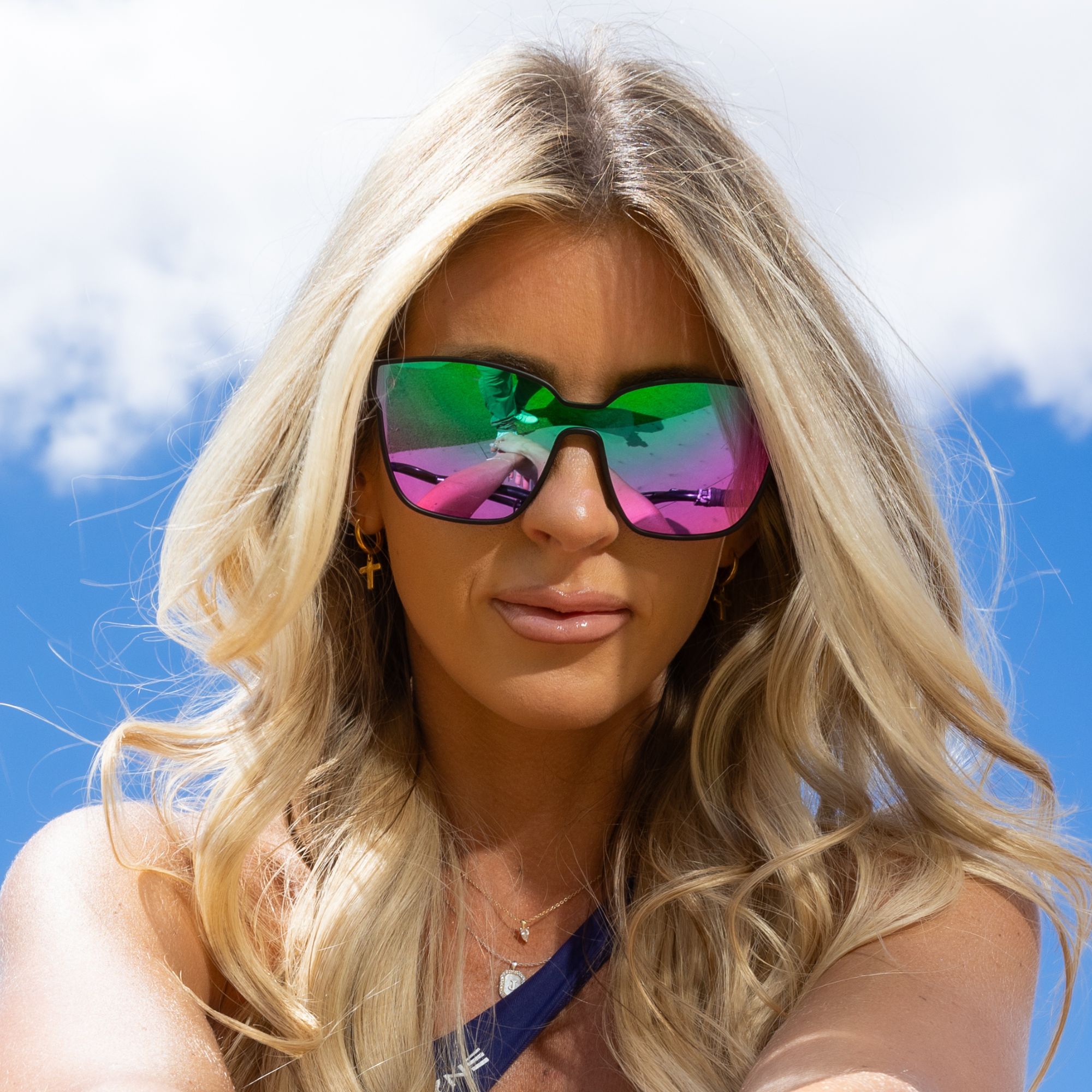 A blonde woman wearing large, reflective green and pink sunglasses against a blue sky with clouds.