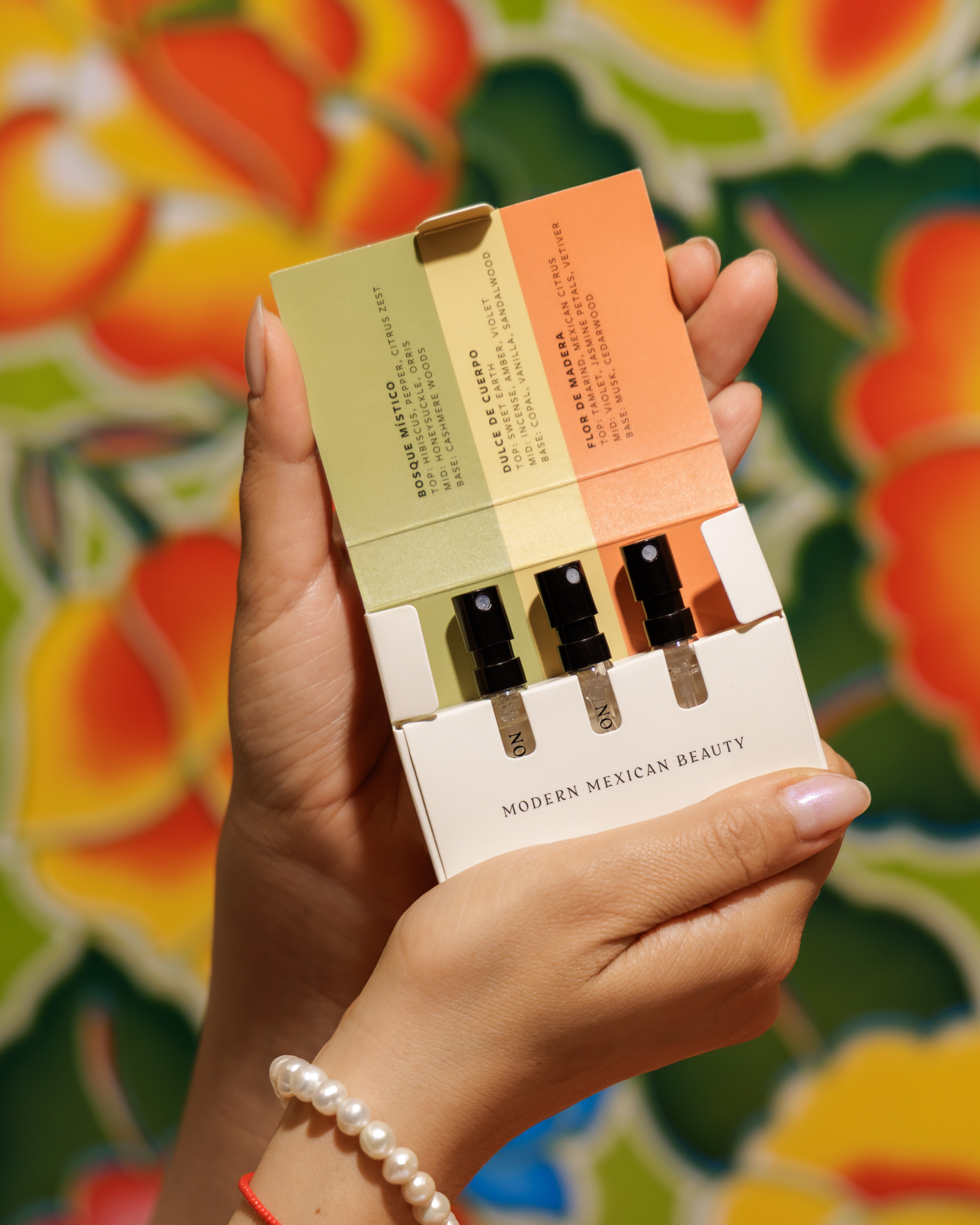 A person's hands holding Nopalera perfumes sampler set with three vials against a colorful floral background.