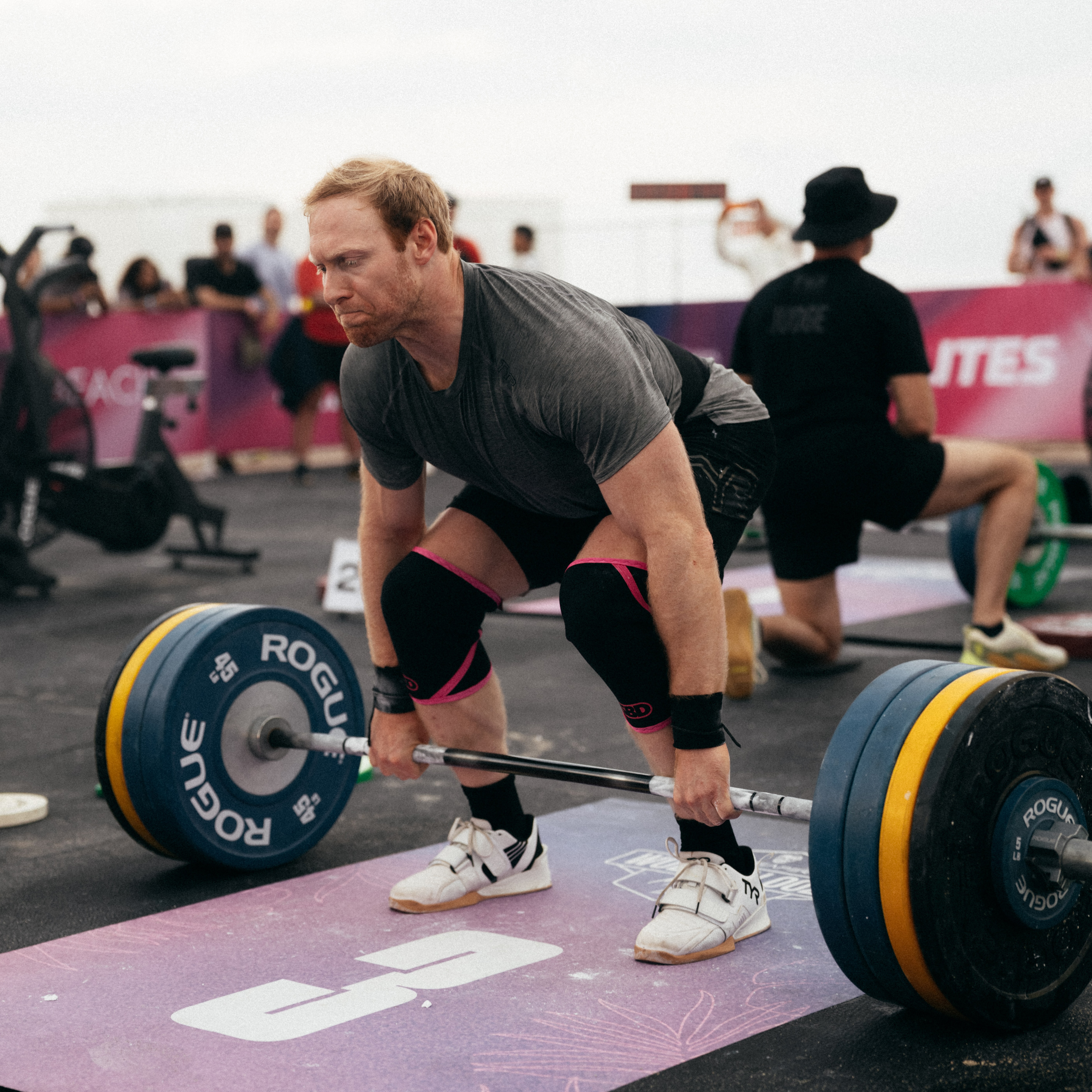 A male weightlifter in a gray shirt bends down to lift a heavy barbell at an outdoor competition.