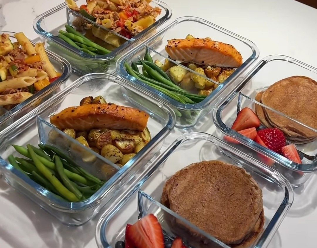 Glass containers with pasta, salmon, green beans, pancakes, and strawberries.