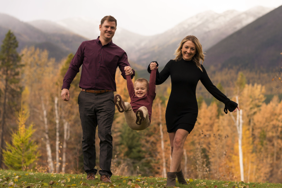 A man and a woman smile as they swing a young child between them outdoors in autumn.