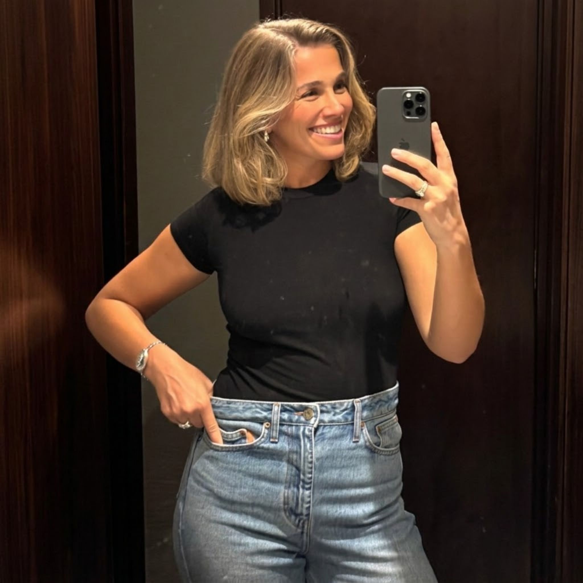 A smiling woman in a black t-shirt and blue jeans takes a mirror selfie with her phone.