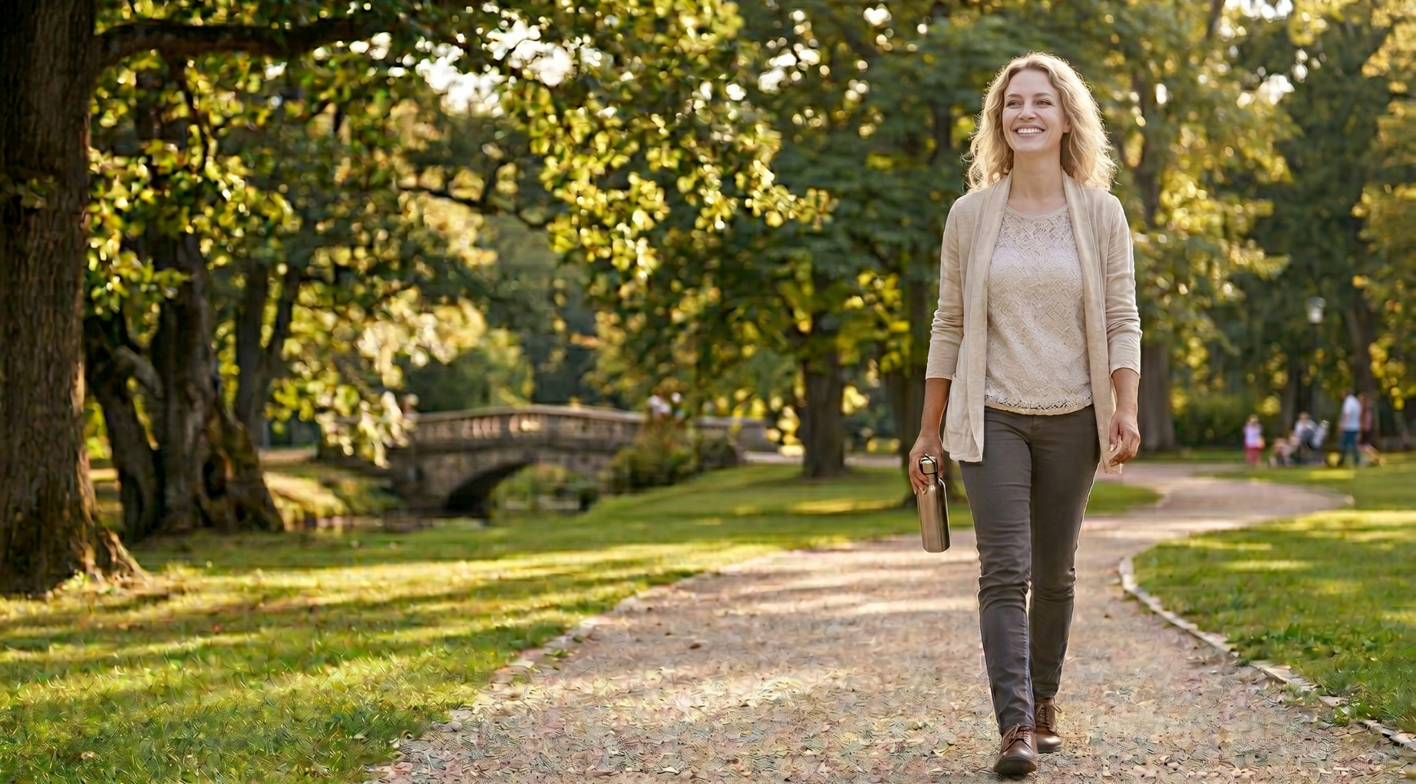 A smiling woman holding a water bottle walks on a path through a sunny park.