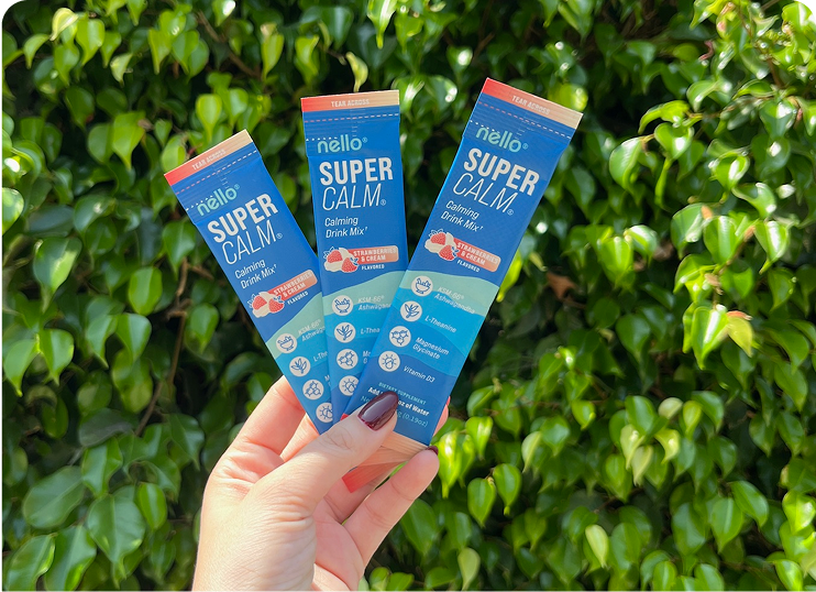 Hand holding three packets of Super Calm drink mix against a leafy background.