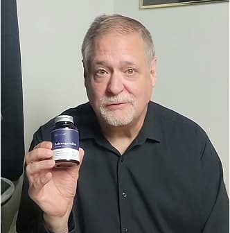 Man holding a bottle with a label, wearing a black shirt, sitting indoors.