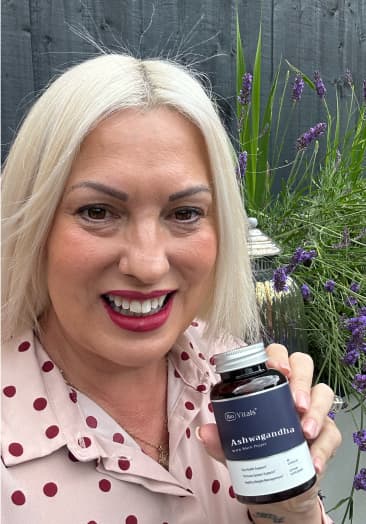 Person holding a bottle of Ashwagandha supplement, smiling outdoors with plants in the background.