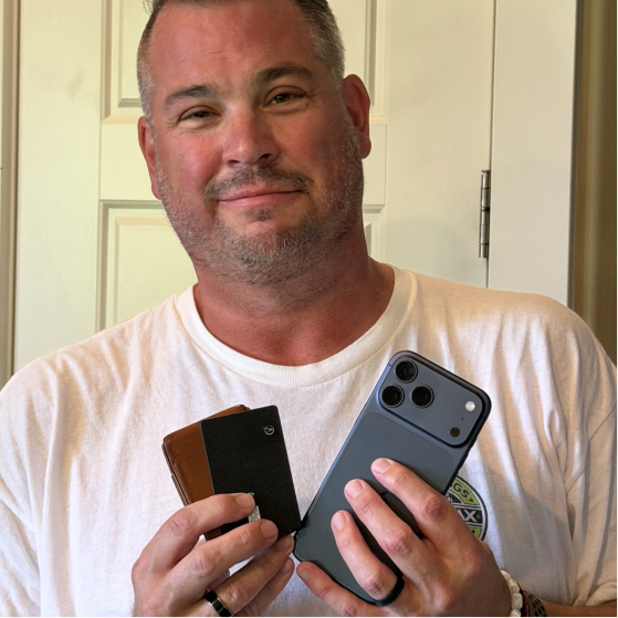 A man in a white t-shirt smiles while holding a smartphone and a wallet.