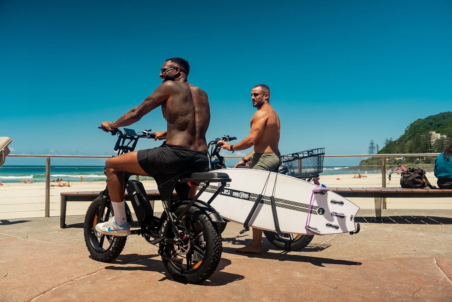 Two shirtless men on electric bikes with a surfboard on a boardwalk next to a sunny beach.